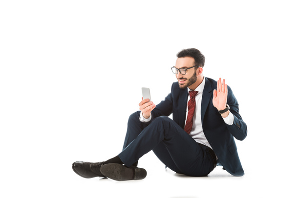 homme d'affaires souriant ayant chat vidéo sur smartphone et agitant la main tout en étant assis sur fond blanc
 - Photo, image