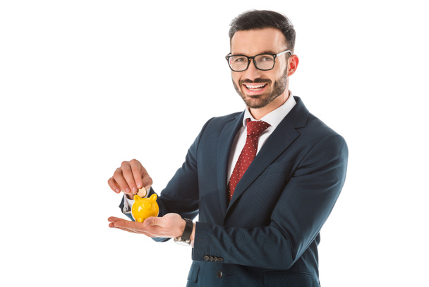 веселый бизнесмен кладет монету в копилку и смотрит на камеру, изолированную на белом
 - Фото, изображение