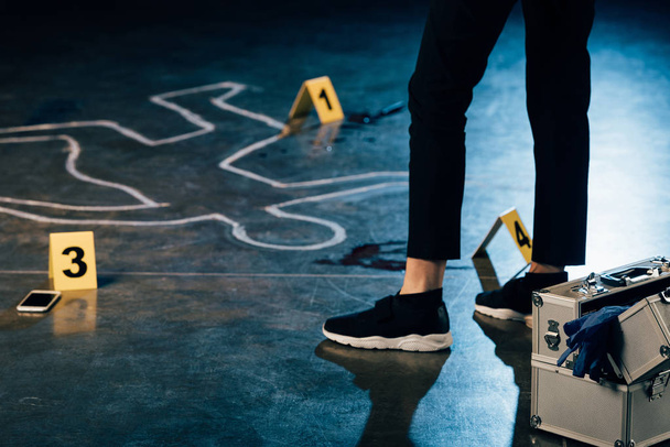 vista parcial del investigador de pie cerca del contorno de tiza y marcadores de evidencia en la escena del crimen
 - Foto, imagen