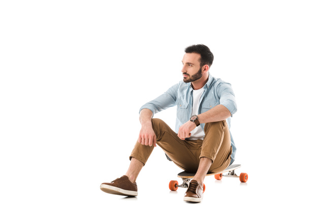 pensive bearded man sitting on longboard and looking away isolated on white - Zdjęcie, obraz