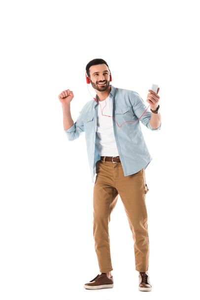 hombre feliz en auriculares escuchando música y sosteniendo el teléfono inteligente mientras baila aislado en blanco
 - Foto, Imagen