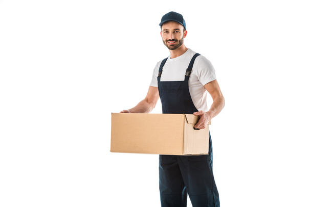 handsome delivery man in overalls holding cardboard box and smiling at camera isolated on white - Фото, зображення
