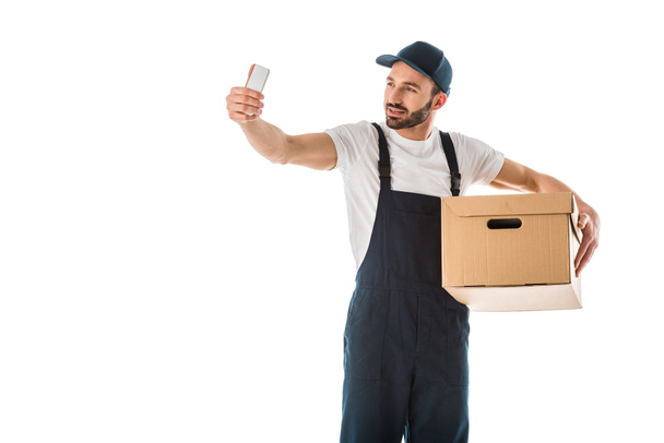 hombre de entrega guapo tomando selfie con teléfono inteligente mientras sostiene la caja de cartón aislado en blanco
 - Foto, imagen