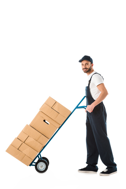 repartidor sonriente transportando camión de mano cargado con cajas de cartón aisladas en blanco
 - Foto, imagen