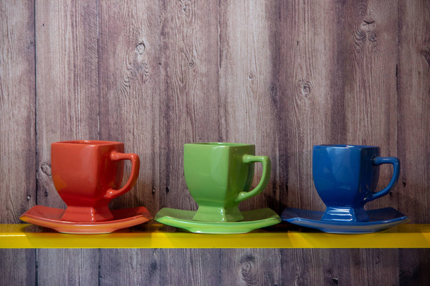 Piros, kék és zöld csészék és csészealjak sárga polcon, fából készült háttéren. - Fotó, kép