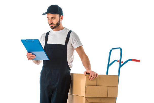 serious delivery man looking at clipboard while standing near hand truck loaded with cardboard boxes isolated on white - Photo, Image