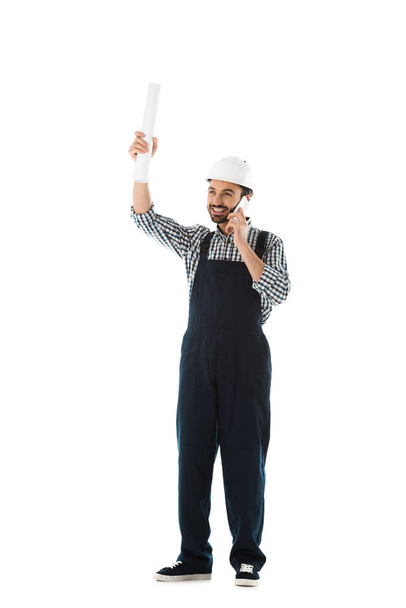 fröhlicher Bauarbeiter, der mit dem Smartphone spricht, während er Papierrollen in der erhobenen Hand hält, isoliert auf weißem Grund - Foto, Bild