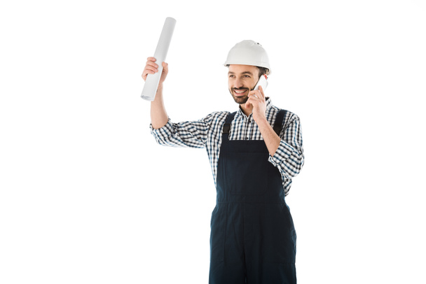 smiling construction worker talking on smartphone while holding rolled paper in raised hand isolated on white - Φωτογραφία, εικόνα