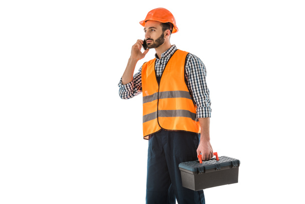 attente bouwvakker die op een smartphone praat terwijl hij Toolbox vasthoudt en op wit kijkt, geïsoleerd - Foto, afbeelding