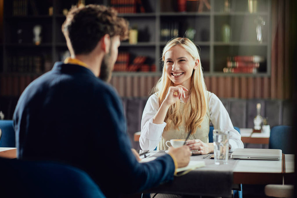 Smiling Caucasian blonde businesswoman dressed smart casual discussing with her male colleague about project while sitting in coffe shop. - Photo, image