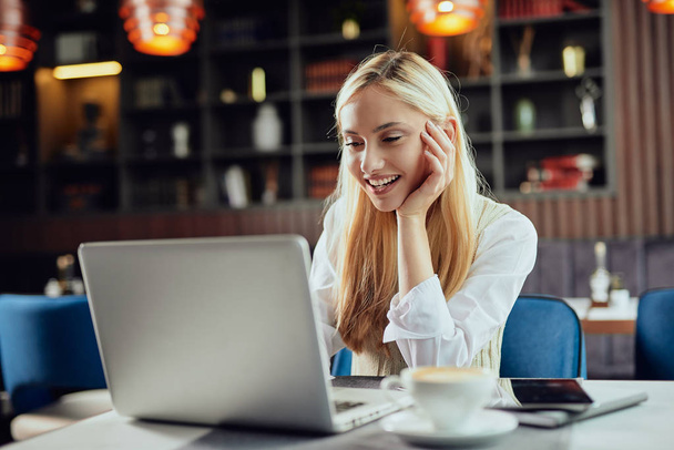 Όμορφη χαμογελαστή Καυκάσιος ξανθός/ιά θηλυκό blogger ντυμένος έξυπνη περιστασιακή κάθεται στην καφετέρια και χρησιμοποιώντας το laptop. - Φωτογραφία, εικόνα