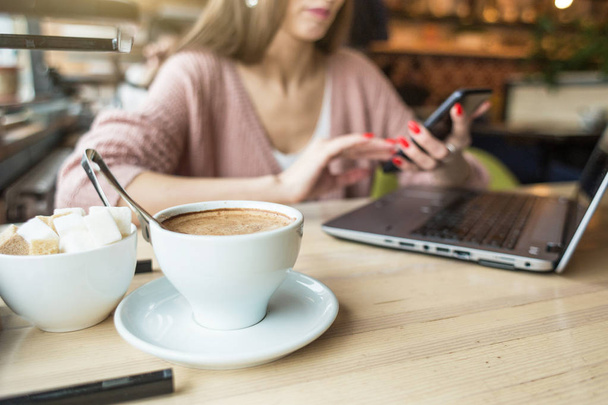Attractive young woman sits at a table in a cafe with a cup of coffee and a laptop and uses the phone - Photo, image