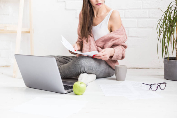 A young happy woman sits on the floor in a bright apartment or office interior and works at a laptop, freelancer girl at work, the concept of distance learning students, paperwork and online learning - 写真・画像