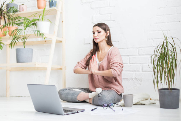 Young happy woman sits on the floor in a yoga pose in a bright apartment and works behind a laptop, a freelancer girl meditates while working. stress relieving - Φωτογραφία, εικόνα