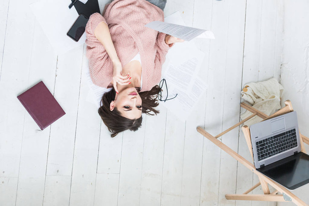 A beautiful woman lies on the floor among papers and documents, the girl freelancer smiles and relaxes during a break from work - Foto, imagen