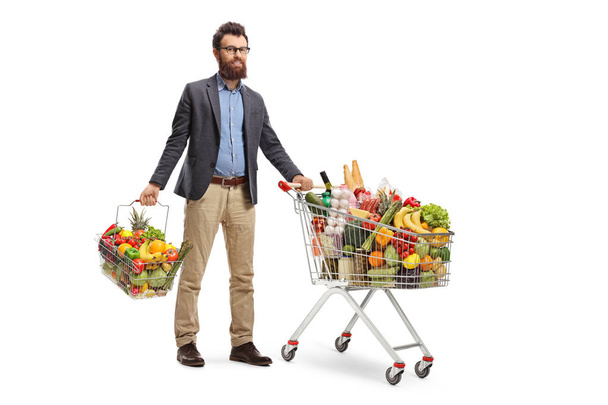 Full length portrait of a young man standing with a shopping cart and a shopping basket with fruits and vegetables isolated on white background - Valokuva, kuva