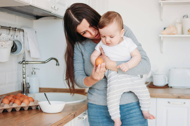 Young mother brunette woman with baby boy in arms cooking food in white modern kitchen at home - Fotografie, Obrázek