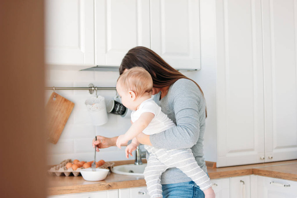 Young mother brunette woman with baby boy in arms cooking food in white modern kitchen at home - Zdjęcie, obraz