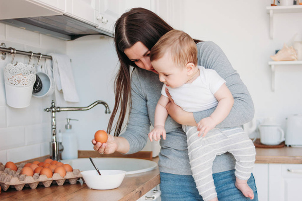 Joven madre morena mujer con bebé niño en brazos cocina comida en blanco cocina moderna en casa
 - Foto, Imagen