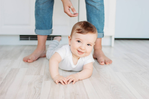 Очаровательный маленький мальчик 6 месяцев улыбается и смотрит в камеру на фоне маминых ног
 - Фото, изображение
