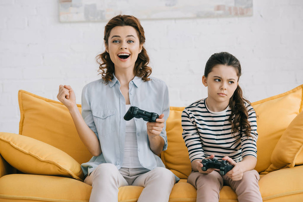 KYIV, UKRAINE - APRIL 8, 2019: Cheerful mother holding joystick and showing yes gesture while sitting near offended daughter - 写真・画像