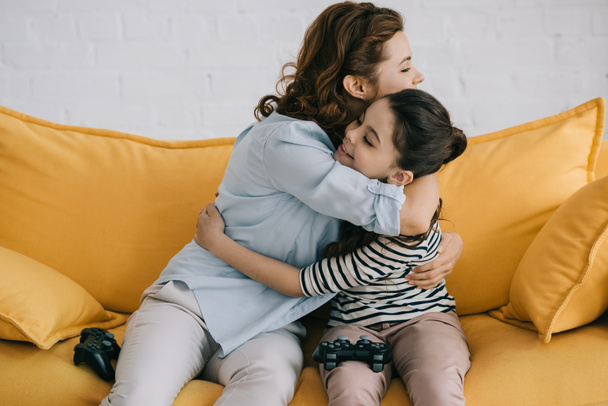 KYIV, UKRAINE - APRIL 8, 2019: Happy mother and daughter hugging while sitting with joysticks on sofa at home - Foto, Imagen