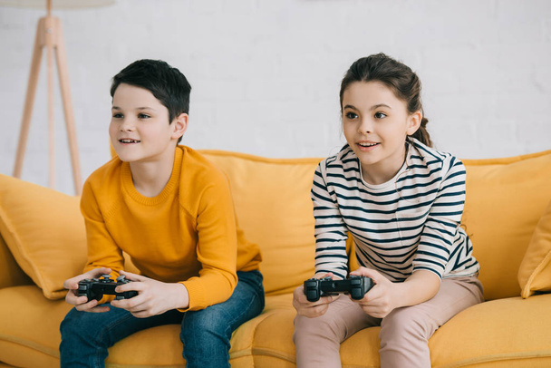 KYIV, UKRAINE - APRIL 8, 2019: Focused brother and sister playing video game with joysticks while sitting on sofa at home - 写真・画像