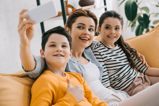 happy attractive woman taking selfie with smartphone while sitting on sofa near smiling daughter and son  - Фото, зображення