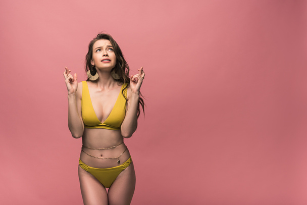 worried girl in yellow swimsuit looking up with crossed fingers isolated on pink - Photo, Image