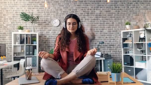 Zoom in time-lapse of woman listening to music in lotus pose on office desk - Video, Çekim
