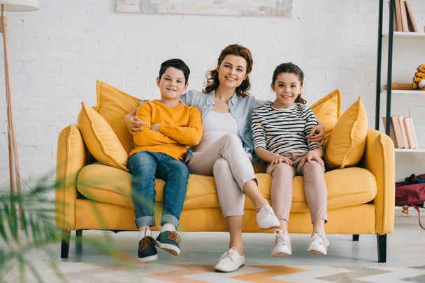Szczęśliwa matka z wesołymi dziećmi siedzi na żółtej kanapie i patrząc na kamerę - Zdjęcie, obraz