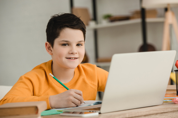 smiling boy writing with pencil and looking at camera while doing schoolwork at home - Foto, imagen