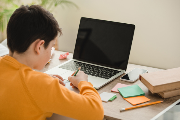 schoolboy doing homework while sitting at desk near laptop with blank screen - Photo, image