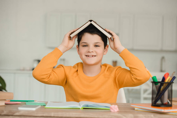 cheerful boy holding open book on head and looking at camera while  - 写真・画像