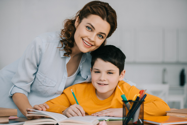 gelukkige moeder met schattige zoon glimlachend en kijkend naar de camera terwijl ze samen huiswerk doen - Foto, afbeelding