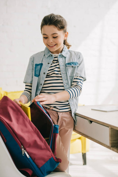 adorable smiling child packing back pack while standing near desk at home - Photo, Image