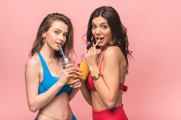 twee mooie meisjes in bikini drinken sinaasappelsap geïsoleerd op roze - Foto, afbeelding