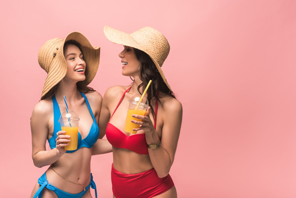 two pretty girls in bikini and straw hats holding disposable cups with orange juice isolated on pink - Foto, Imagem