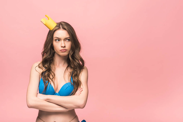Unzufriedenes Mädchen im Bikini und Krone posiert mit verschränkten Armen auf rosa - Foto, Bild