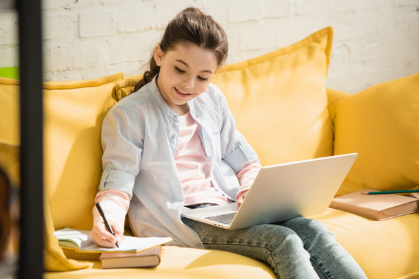 χαμογελαστός προσεκτικός παιδί γράφοντας στο βιβλίο αντιγραφής και χρησιμοποιώντας το laptop, ενώ κάθονται στον καναπέ στο σπίτι - Φωτογραφία, εικόνα