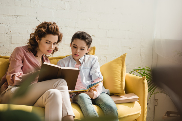 mother and daughter sitting on yellow sofa at home and reading book together - Photo, Image