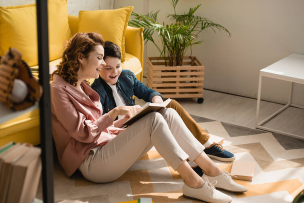 cheerful mother and son sitting on floor at home and reading book together - Photo, image