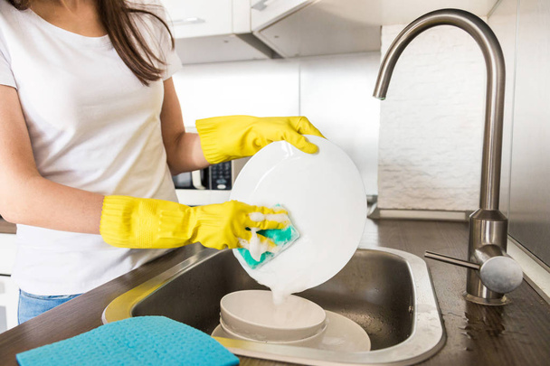 Egy fiatal nő sárga kesztyűben mosogatja a tányérokat szivaccsal a mosogatóban. Háztartási professzionális takarítás. - Fotó, kép