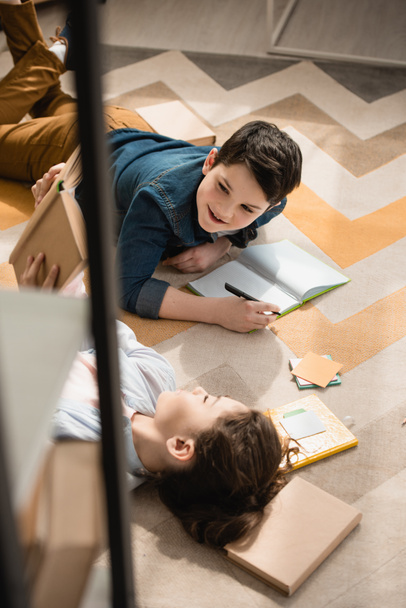 high angle view of cute brother and sister lying on floor and doing homework - Photo, Image