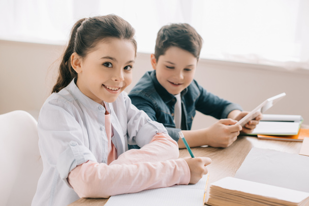 selektiver Fokus des lächelnden Kindes, das in Notizbuch schreibt und in die Kamera schaut, während es mit seinem fröhlichen Bruder Hausaufgaben macht  - Foto, Bild