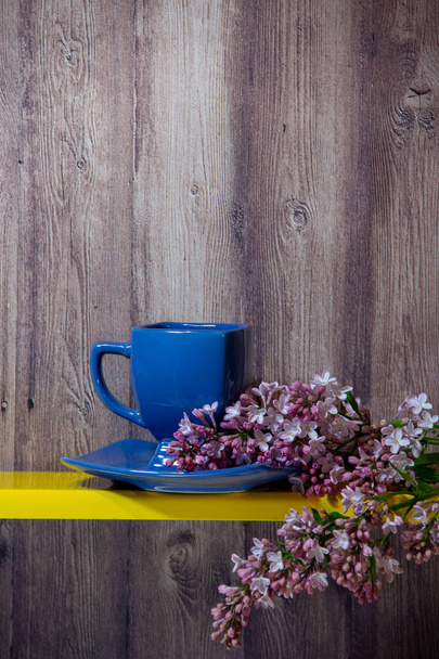Ένα μπλε φλιτζάνι, πιατάκι και κλαδί μιας άνθιας άνοιξης λιλά Stand σε ένα κίτρινο ράφι, πίσω είναι ένα ξύλινο φόντο. - Φωτογραφία, εικόνα