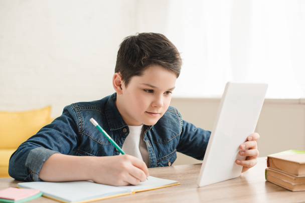 концентрированный мальчик пишет в блокноте и с помощью цифрового стола, выполняя школьные задания на дому
  - Фото, изображение