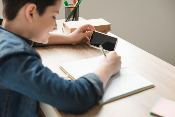 selective focus of boy writing in notebook and holding smartphone with blank screen - Photo, Image