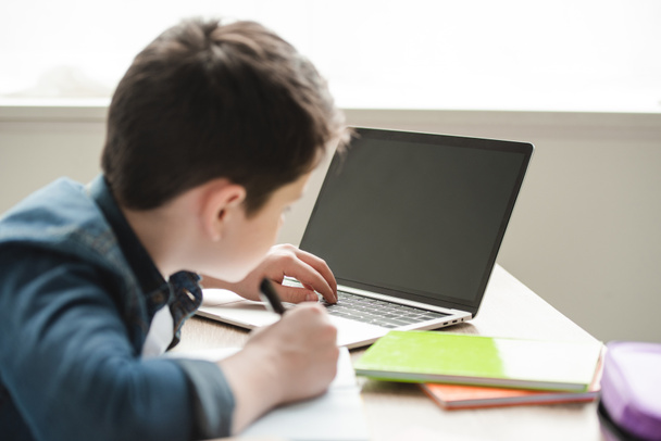 μαθητής γράφοντας στο σημειωματάριο και χρησιμοποιώντας το laptop, ενώ κάνει σχολικές εργασίες στο σπίτι - Φωτογραφία, εικόνα