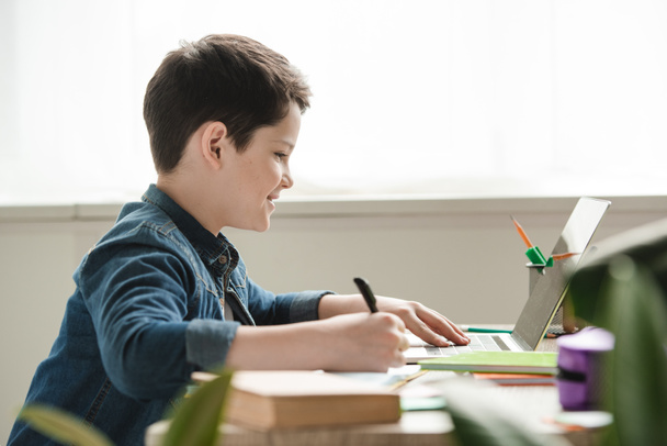 lachende jongen schrijven in notebook en het gebruik van laptop terwijl het doen van school werk thuis - Foto, afbeelding
